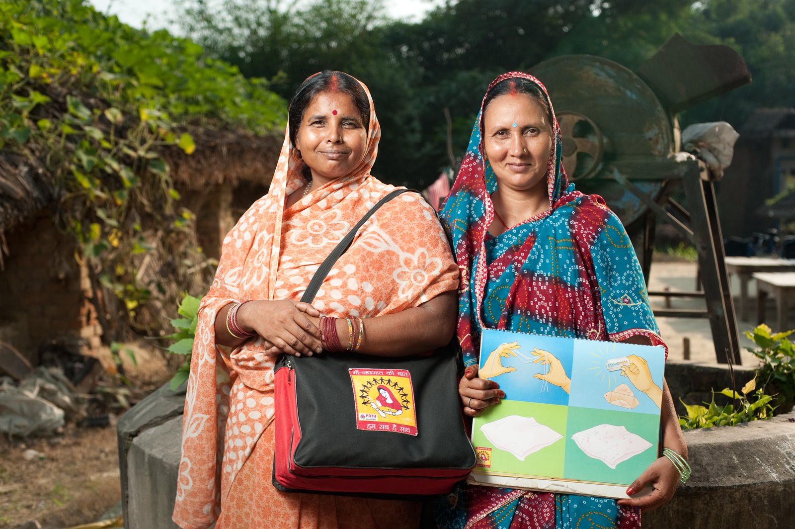 Two women in saris smile for the camera, one holding an over-the-shoulder bag with health supplies and the other holding an instructional poster.