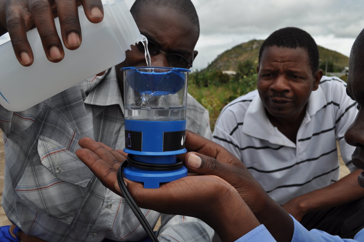 A man pours water into an MSR Community Chlorine Maker (electrochlorinator) while another man holds the device and a third man looks on.