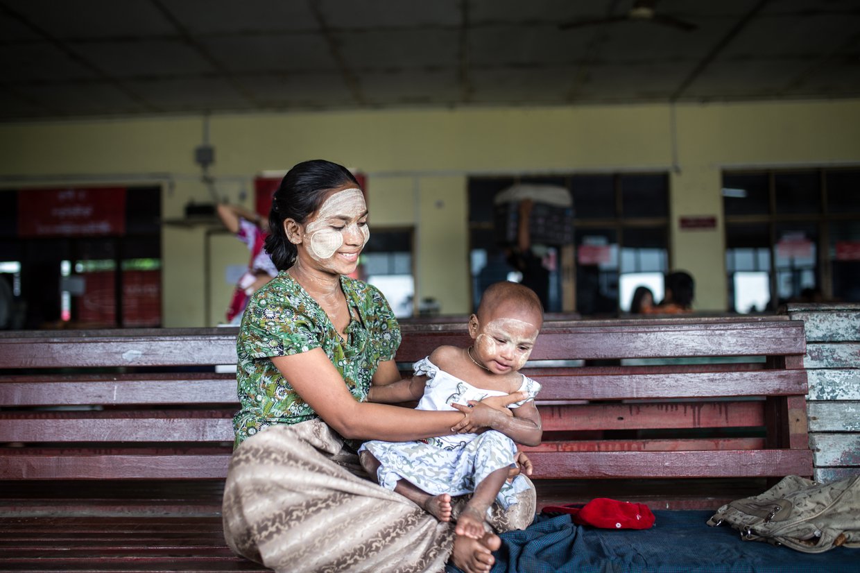 A Burmese woman with Thanakha paste on her face holds a toddler while sitting on a bench.