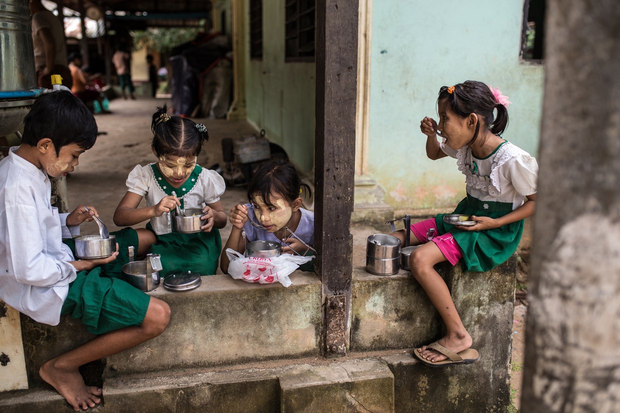 Four children sit in a doorway eating from metal containers.