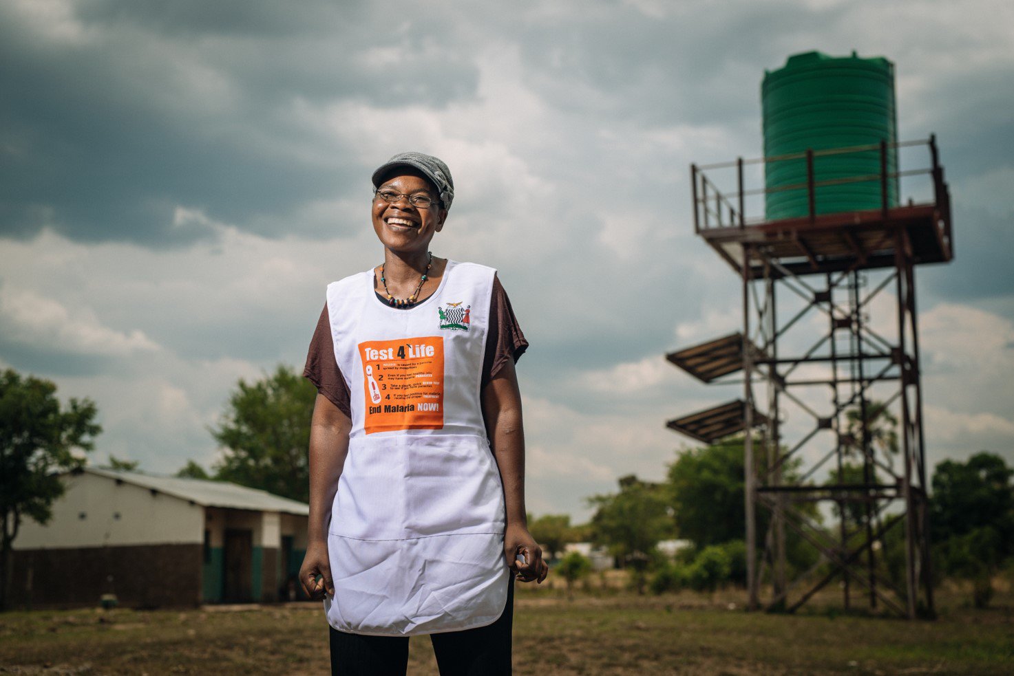 recious Namakau, community health worker, 38 years old, 3 children, wearing cap and glasses, test and treat apron, stands outside with the Mandia community health post's water tower in the background. Photo: PATH/Gabe Bienczycki